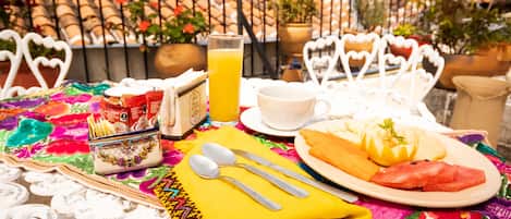 Una mesa dispuesta con alimentos para el desayuno, incluyendo un plato de comida, un vaso de zumo de naranja y una taza, todo bajo un paraguas decorativo.
