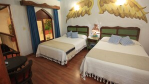 A room with two beds, wooden flooring, a nightstand with a lamp, and a window with a view of greenery.