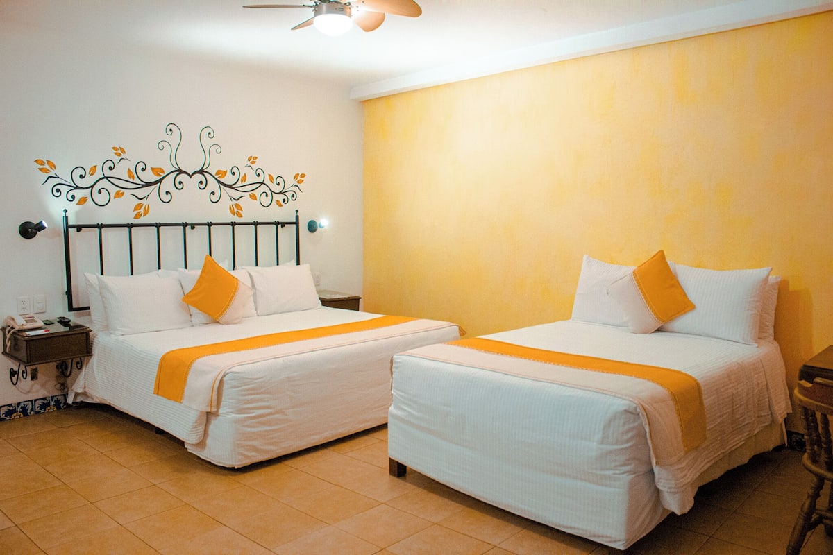 A hotel room with two beds, each with white bedding and orange accents, a decorative wall headboard, and a ceiling fan.