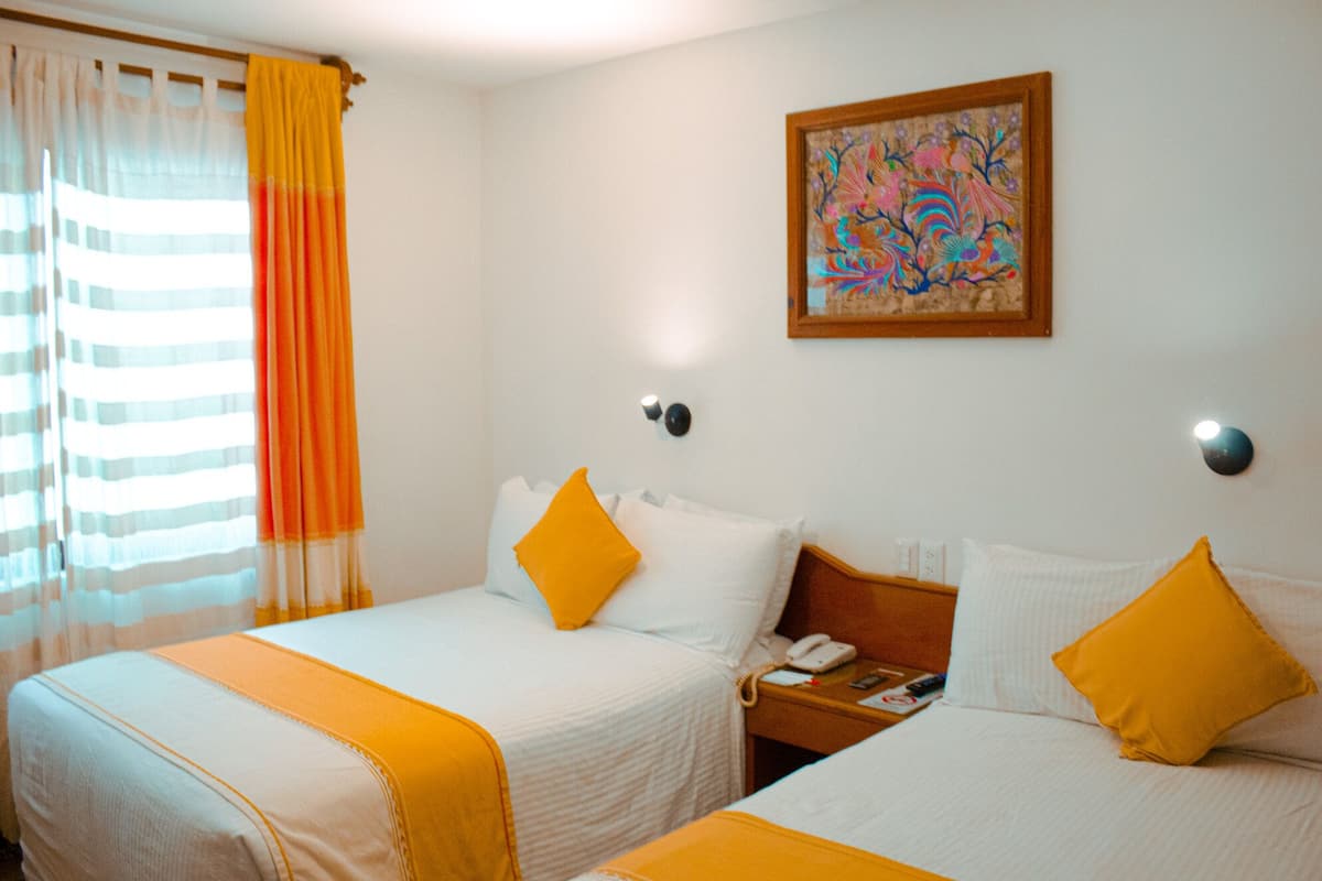 A hotel room with two beds, each with a white bedspread and a yellow throw. There is a painting on the wall above the beds.
