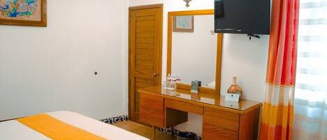 Quarto de hotel com cama, penteadeira de madeira, espelho, televisão e janela com cortinas.