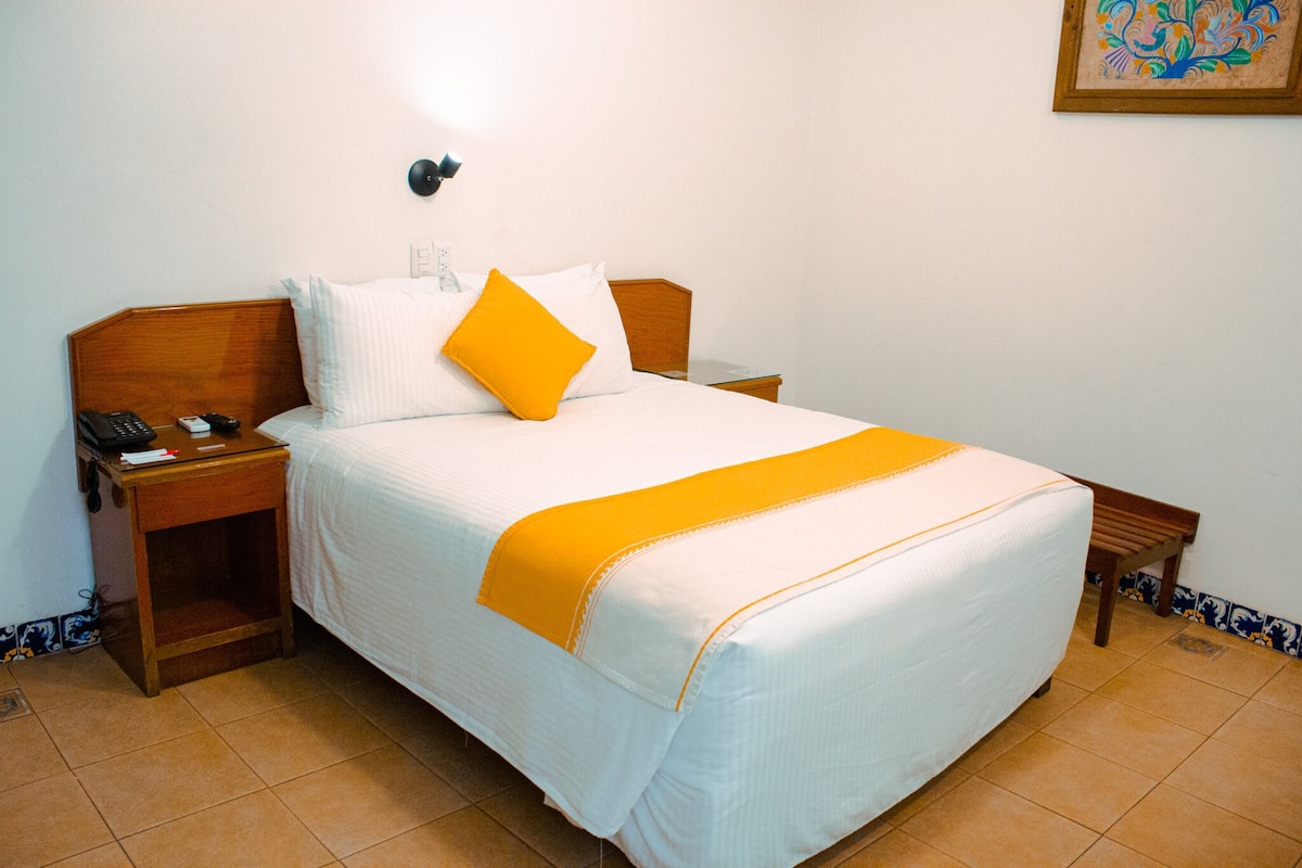 A neatly made bed with white bedding and a yellow accent pillow, a bedside table with a phone and a lamp, and a small wooden bench beside the bed.