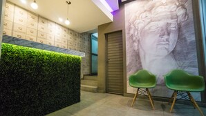 Un área de recepción con una pared verde, piso de baldosas de mármol y un mural grande en la pared. Hay dos sillas verdes y una puerta al fondo.