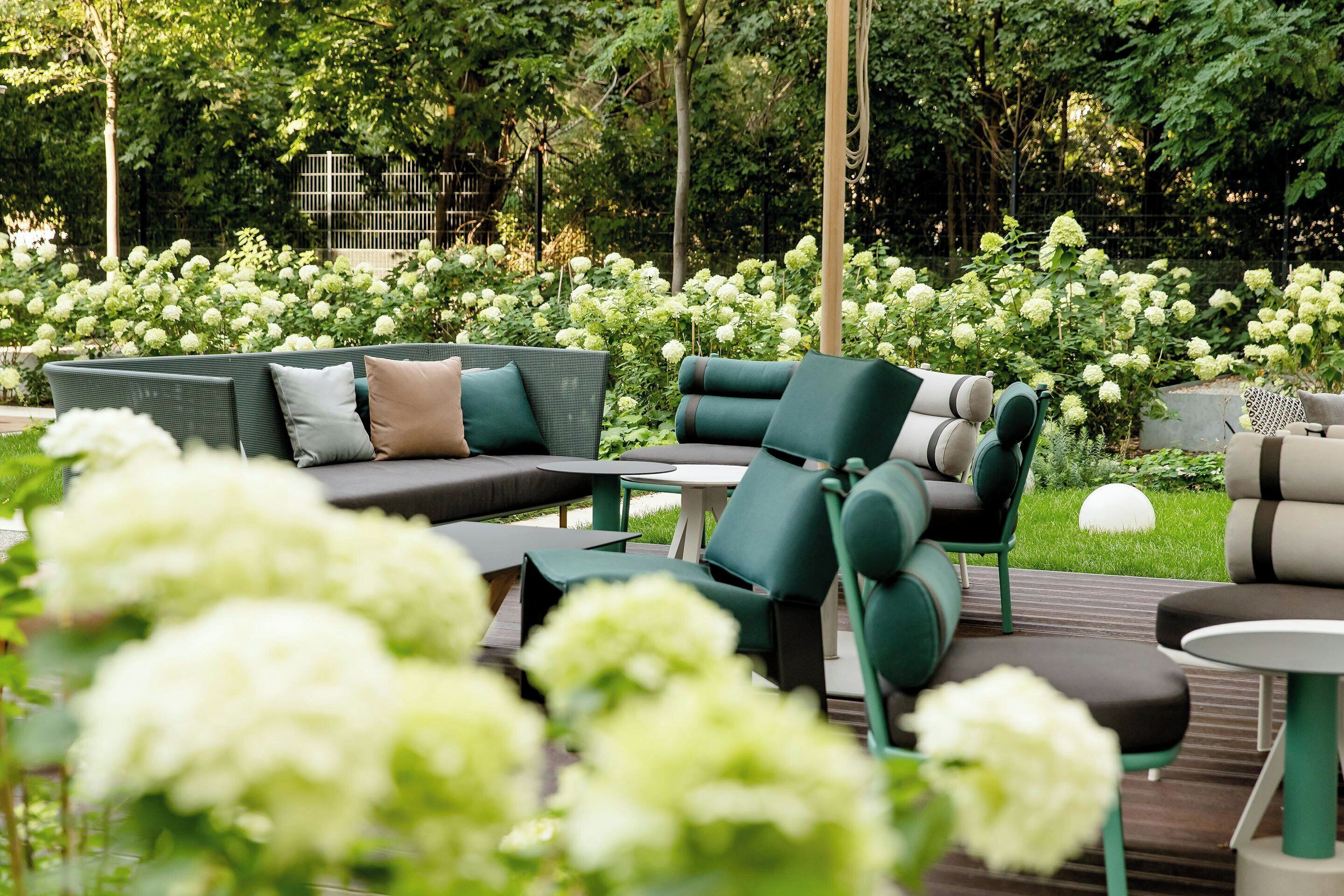 An outdoor seating area with green sofas and chairs, surrounded by lush greenery and white flowers.