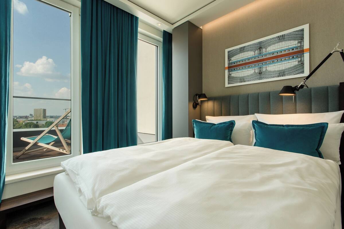Quarto de hotel com uma cama grande, vista da cidade pela janela, uma cadeira junto à janela e um quadro na parede.