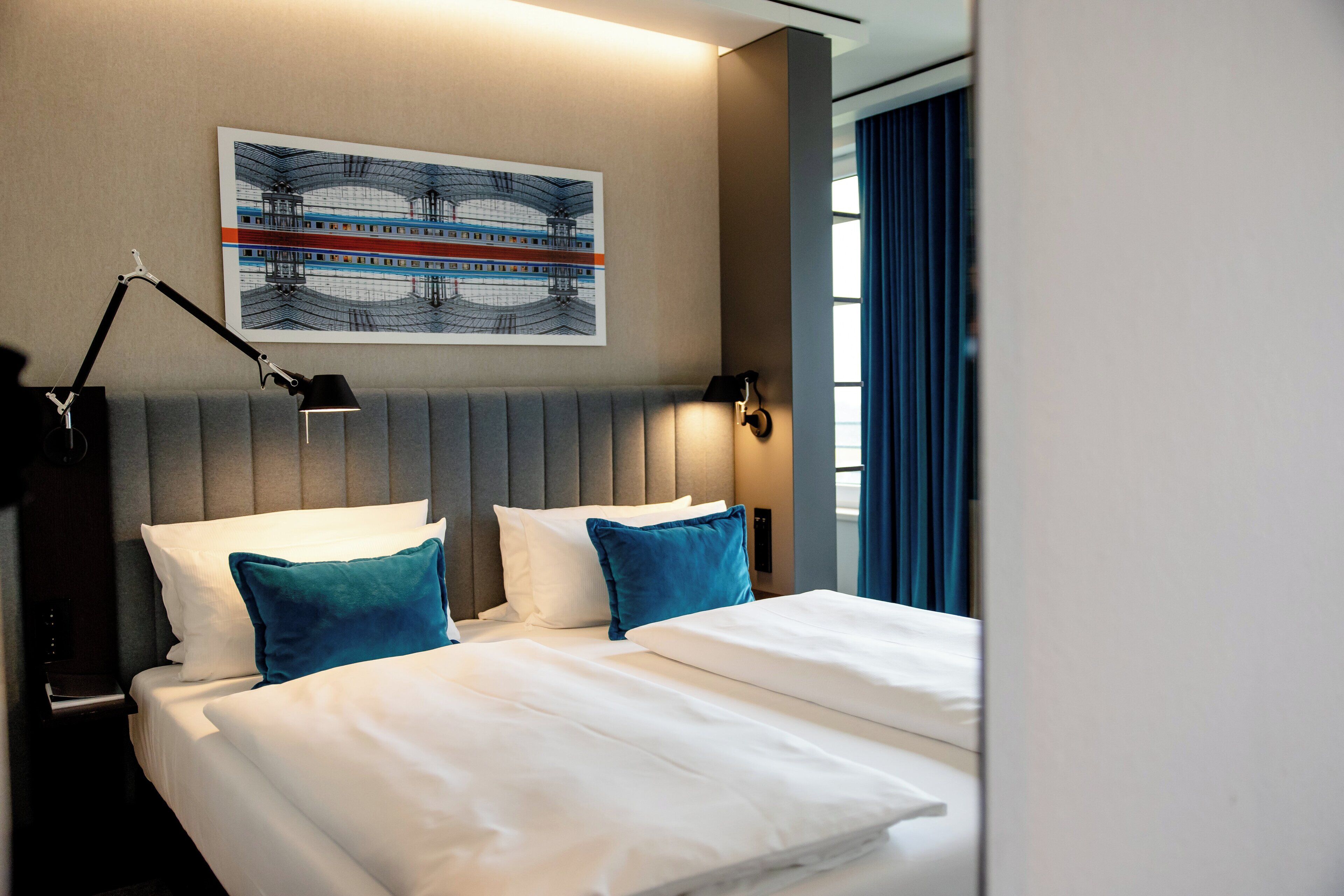 Ein Hotelzimmer mit Bett, Nachttisch, Lampe, Wandbild und einem Fenster mit Vorhängen.