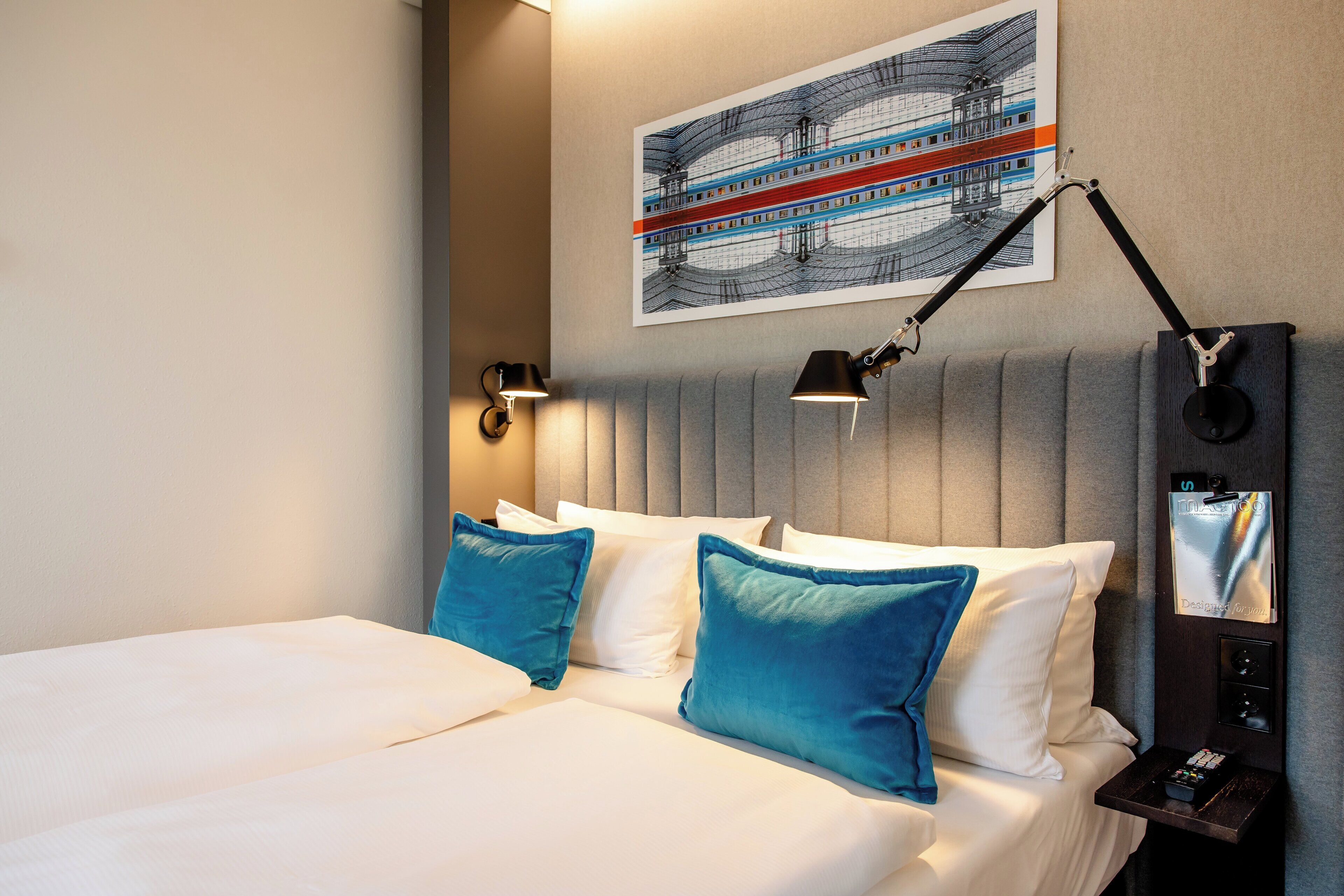 A hotel room with a bed, two pillows, a headboard, a wall-mounted artwork, two bedside lamps, and a nightstand with a remote control.