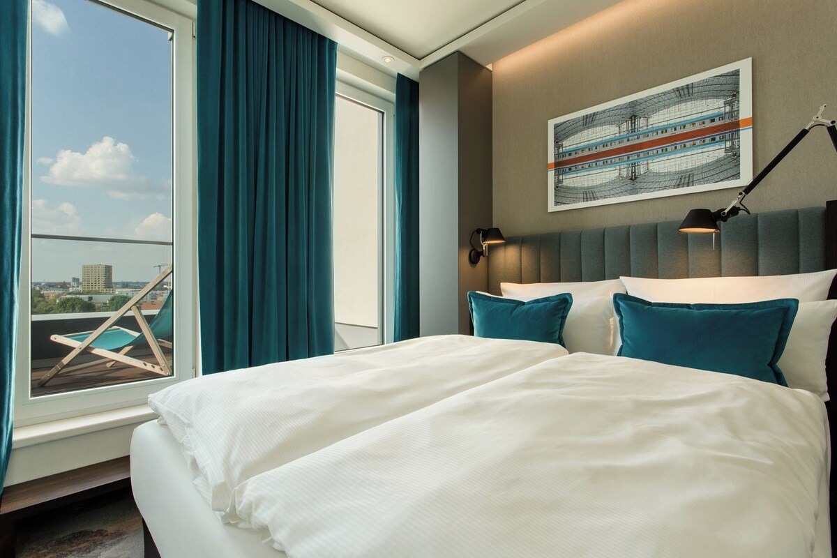 Camera d'albergo con un letto grande, vista sulla città attraverso una finestra, una poltrona vicino alla finestra e un quadro appeso al muro.