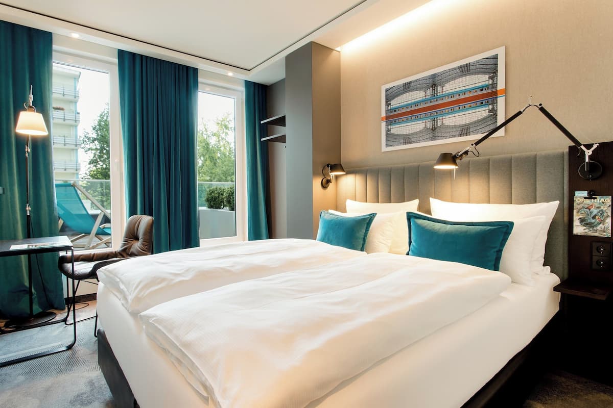 Quarto de hotel com uma cama grande, uma escrivaninha e uma cadeira. Há uma janela com cortinas, uma luminária na parede e um quadro sobre a cama.
