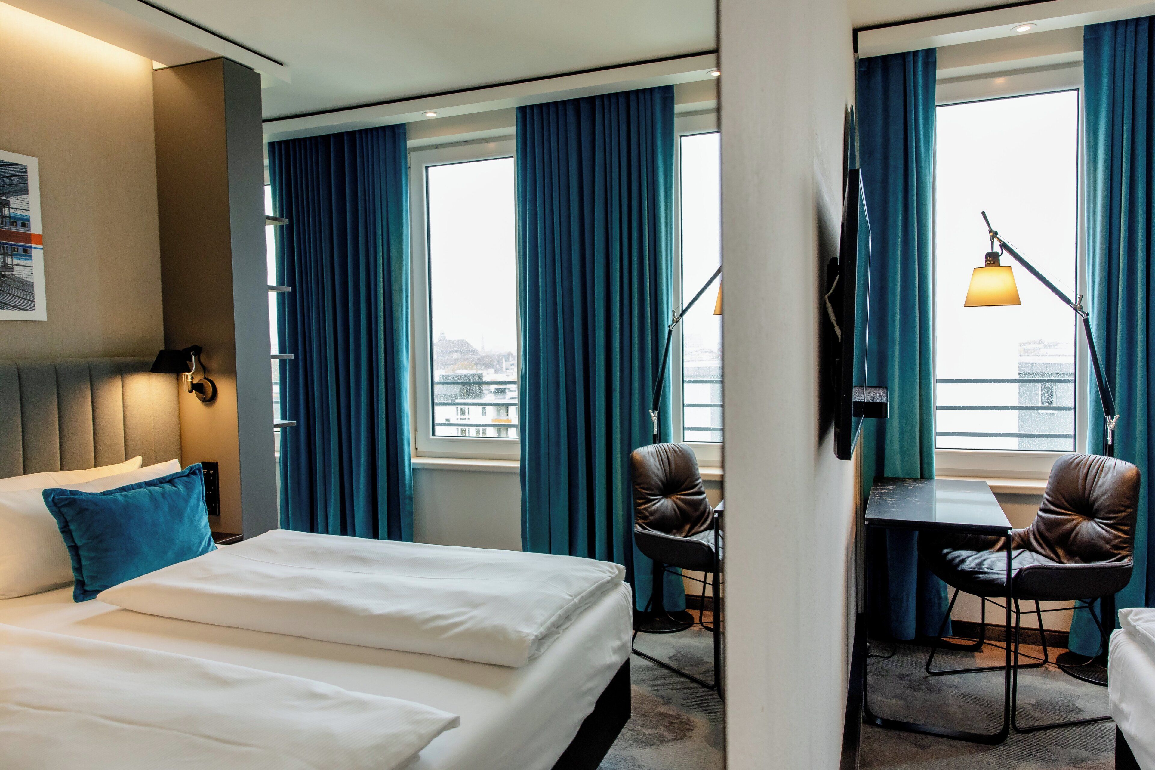 Une chambre d’hôtel avec un grand lit, deux fauteuils, un bureau et une vue sur la ville grâce à de grandes fenêtres.