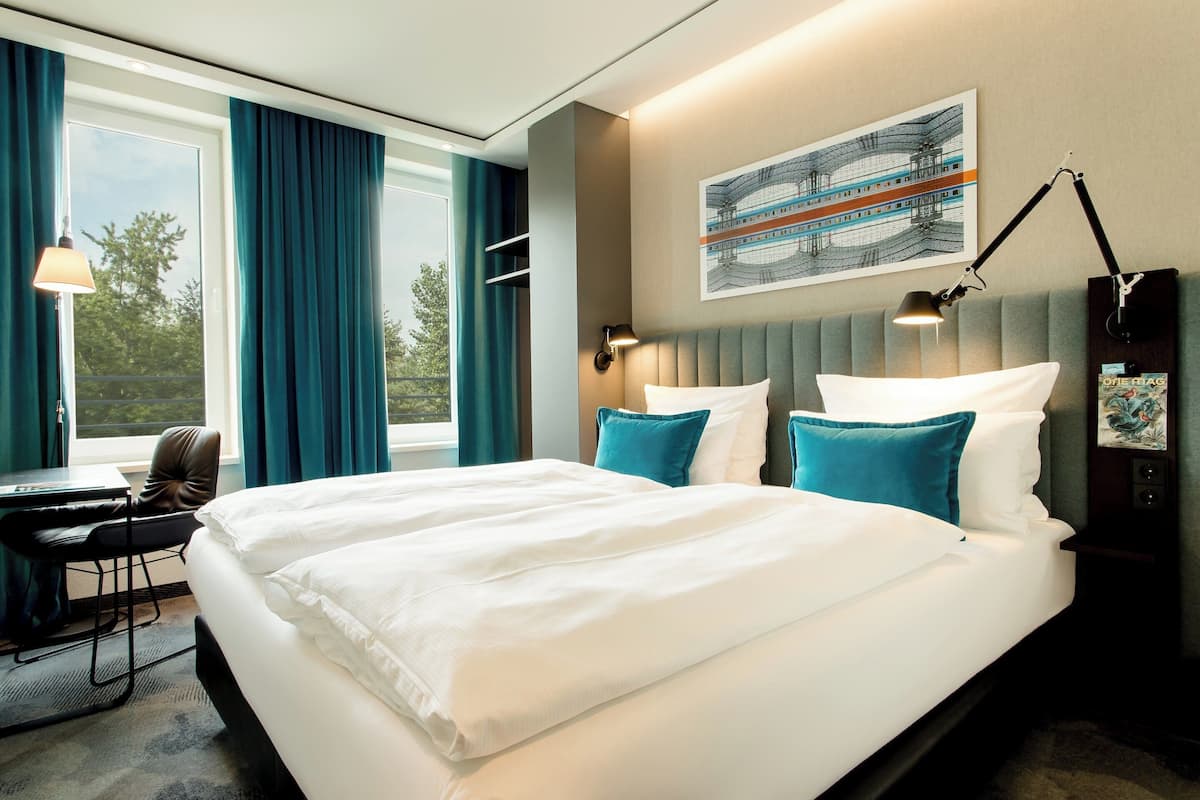 Quarto de hotel com uma cama grande, uma escrivaninha e uma cadeira. Há uma janela com cortinas, um quadro na parede e um abajur na mesa de cabeceira.