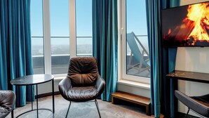 A modern room with a large window, a leather chair, a small table, and a flat-screen TV displaying a fire.