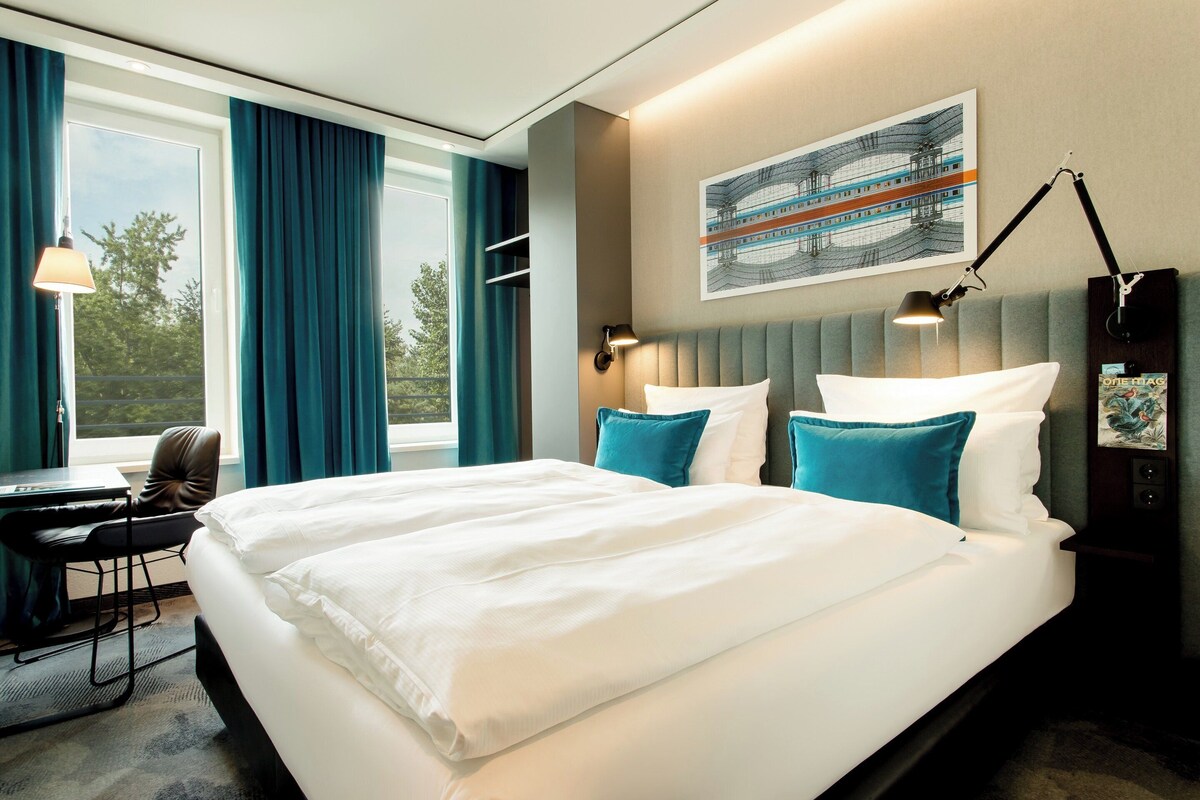 Camera d'albergo con un letto grande, una scrivania e una sedia. Si vede una finestra con tende, un quadro appeso al muro e una lampada sul comodino.
