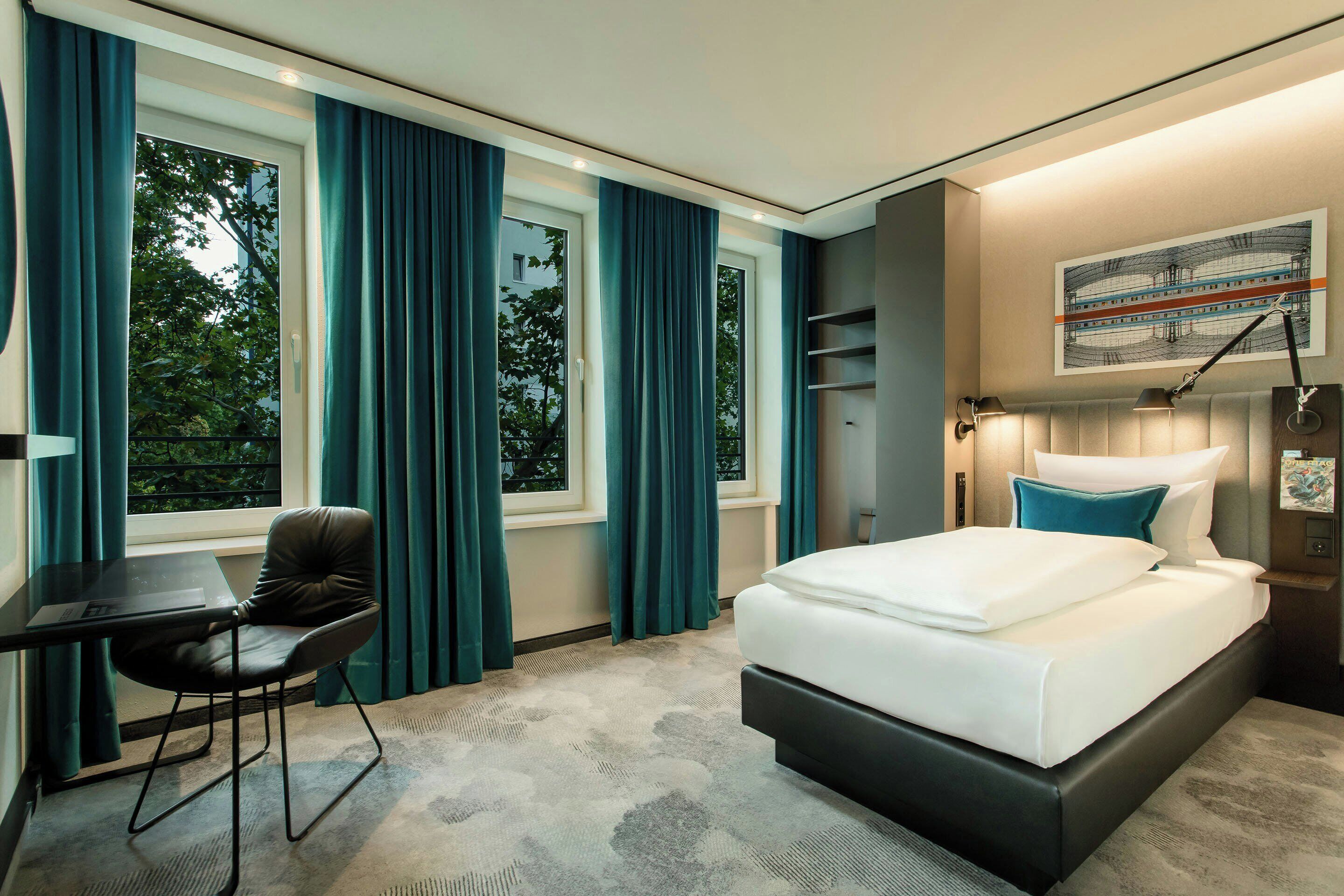 A hotel room with a bed, a desk, and a chair. There are curtains on the windows, and a painting on the wall.