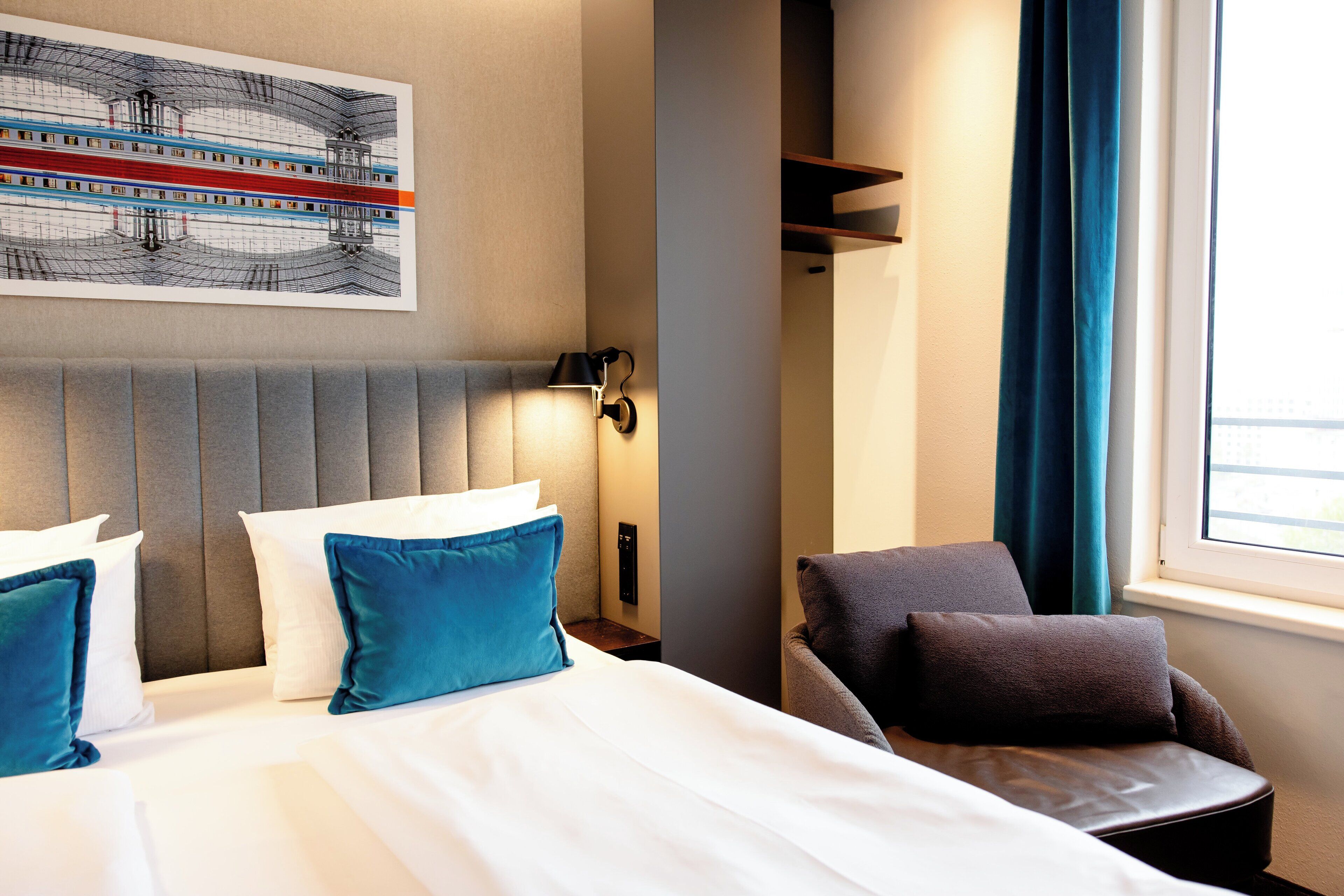 A hotel room with a bed, a chair, a wall-mounted lamp, and a window with curtains.