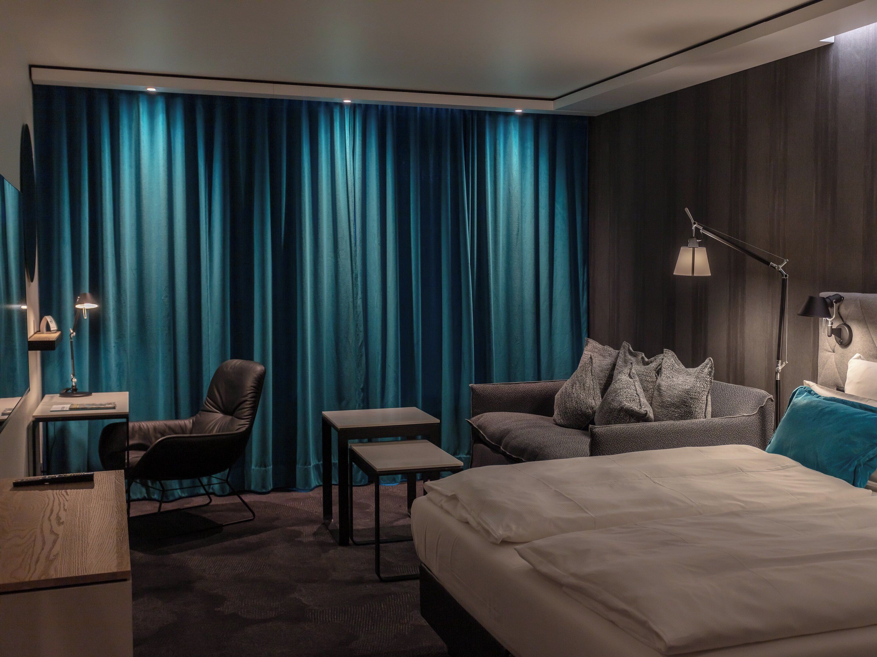 Habitación de hotel con una cama grande, un sofá, un escritorio y una silla. Hay dos lámparas y una cortina.