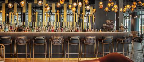 A bar area with multiple hanging spherical light fixtures, a variety of bottles on the bar counter, and several seating options including patterned chairs and a green ottoman.