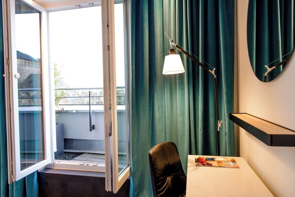 A hotel room with a view through an open door, a desk with a chair, a lamp, and a magazine on the table.
