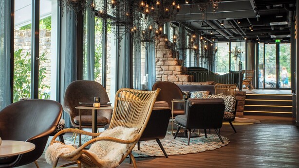 A modern interior with wicker furniture, a stone fireplace, and large windows offering natural light. - Motel One Saarbrücken (Saarbrücken)