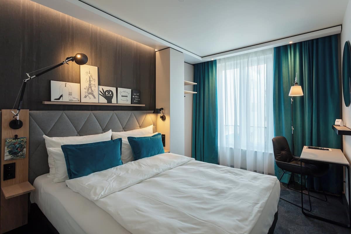 A hotel room with a large bed, a desk, a chair, and a desk lamp.