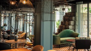 A cozy cafe with a wooden table, chairs, and a green sofa. There are hanging lights and a stone wall in the background. - Motel One Saarbrücken (Saarbrücken)