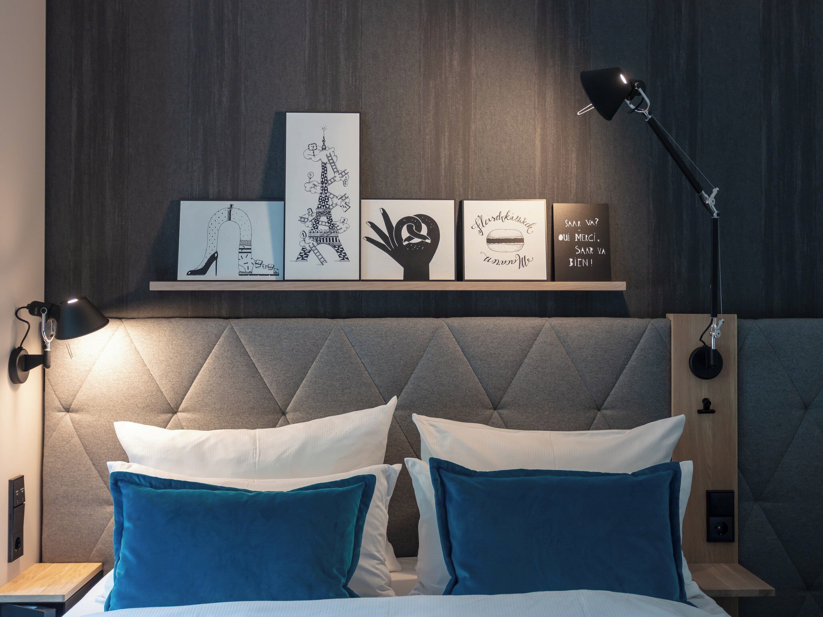 Quarto moderno com cabeceira estofada, cama com almofadas brancas e azul-turquesa, mesa de cabeceira, luminária de parede e prateleira com obras de arte emolduradas.