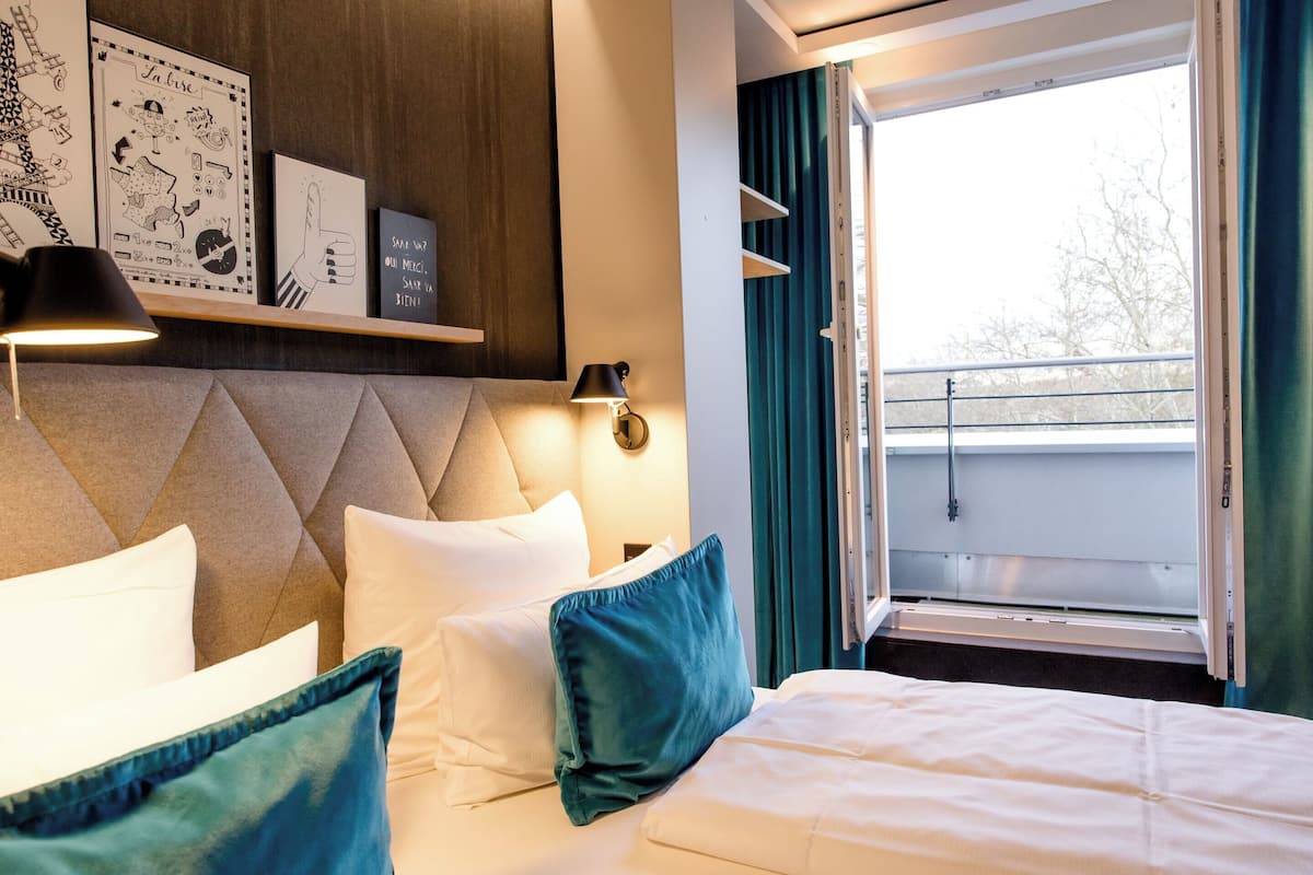 A hotel room with a bed, pillows, a headboard, a wall-mounted shelf with artwork, a window with curtains, and a balcony view.
