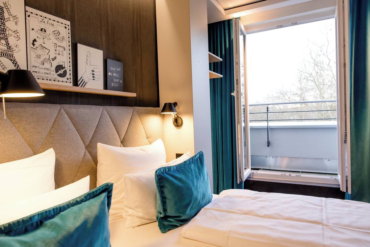 Habitación de hotel con cama, almohadas, cabecero, repisa con obras de arte y balcón con vistas.