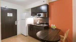 A compact kitchen with dark cabinets, a microwave, a refrigerator, and a round dining table with a bowl of fruit. - Motel 6 Brandon, MB (Brandon)