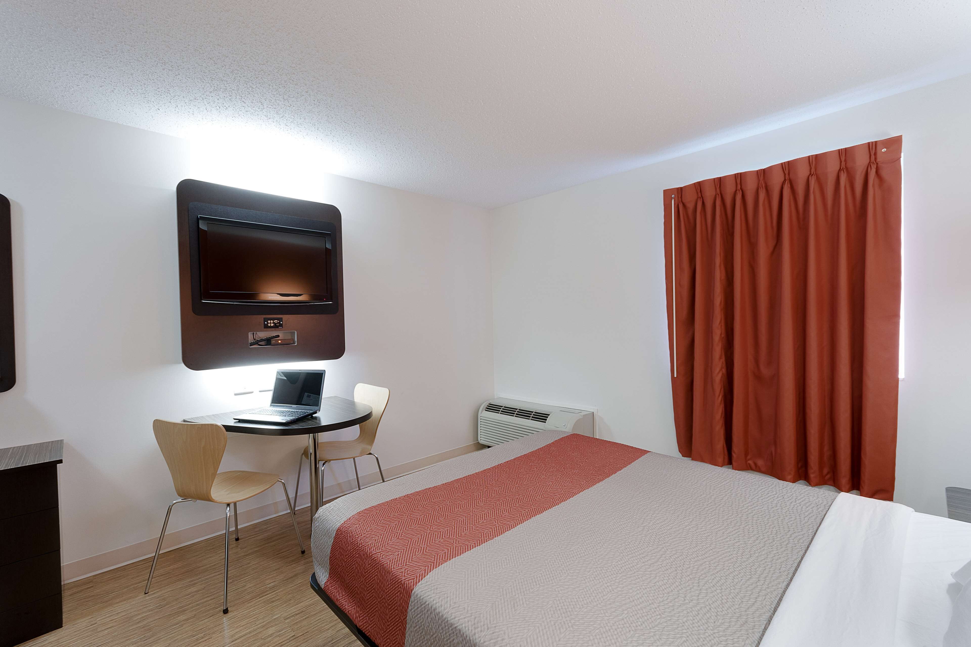 Una habitación de hotel con una cama, un escritorio con una computadora portátil, una silla, un televisor montado en la pared y una ventana con cortinas.