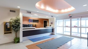 A reception area with a counter, a plant, a blue welcome mat, and a large glass door leading outside. - Motel 6 Brandon, MB (Brandon)