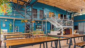 Amplio espacio interior con mesas y bancos de madera, una escalera y una pared azul con un mural.