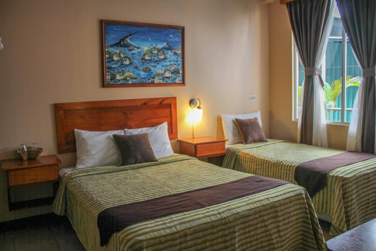 Habitación de hotel con dos camas, una mesita de noche, una lámpara, un cuadro en la pared y una ventana con cortinas.