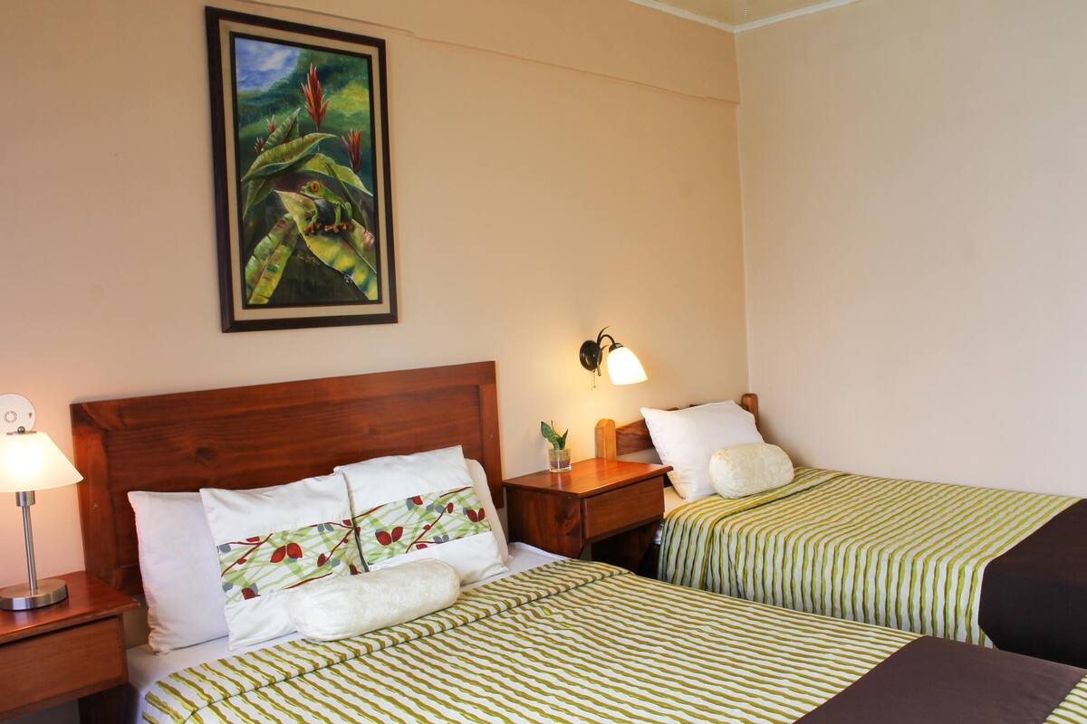 Habitación de hotel con dos camas, una mesita de noche con lámpara, un cuadro enmarcado en la pared y una planta en la mesita de noche.