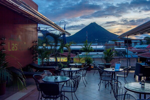 An outdoor patio with tables and chairs, overlooking a mountain and a body of water. - Hotel Las Colinas (La Fortuna)