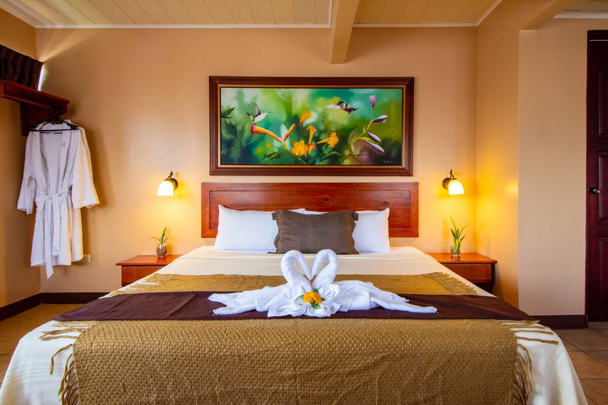 Habitación de hotel con una cama grande, un cuadro sobre la cama, dos lámparas de noche, una bata colgada en un perchero y una toalla dispuesta artísticamente en la cama.