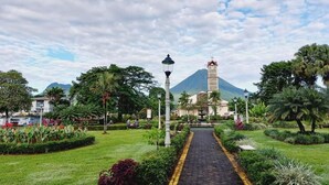 Un sendero conduce a un edificio con torre, rodeado de abundante vegetación y una montaña al fondo.