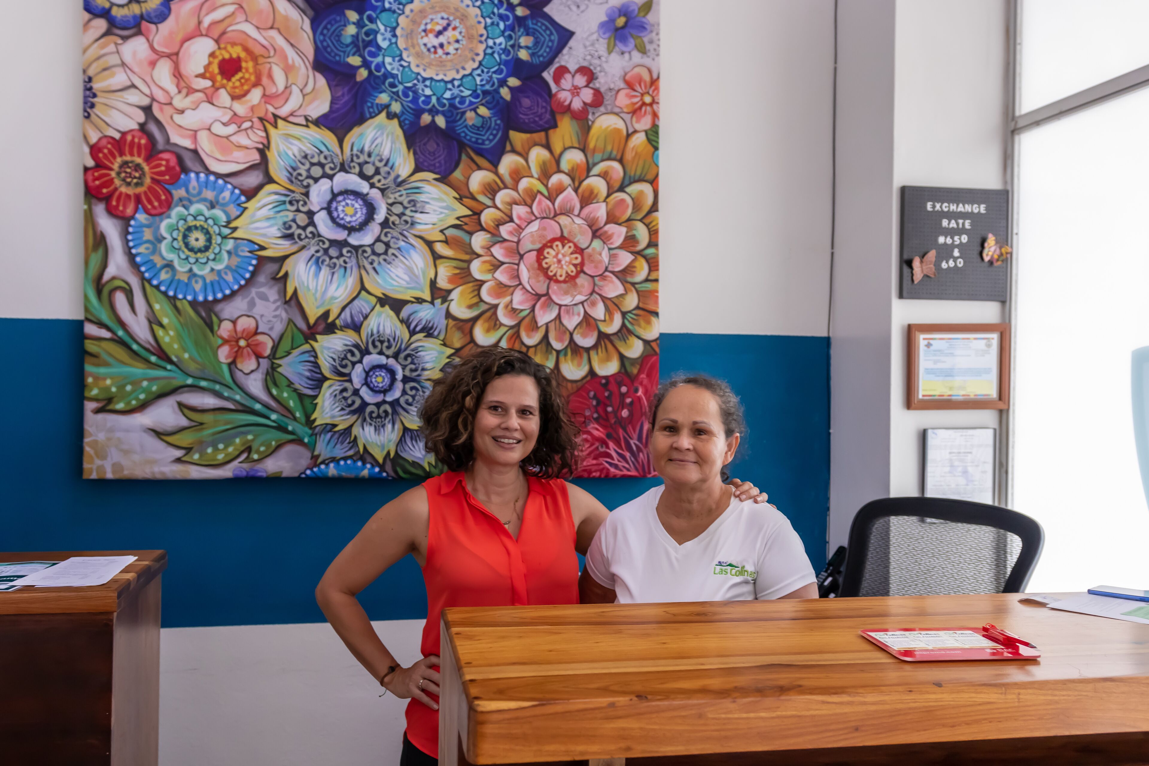 Two people standing at a wooden table with a colorful floral painting on the wall behind them.
