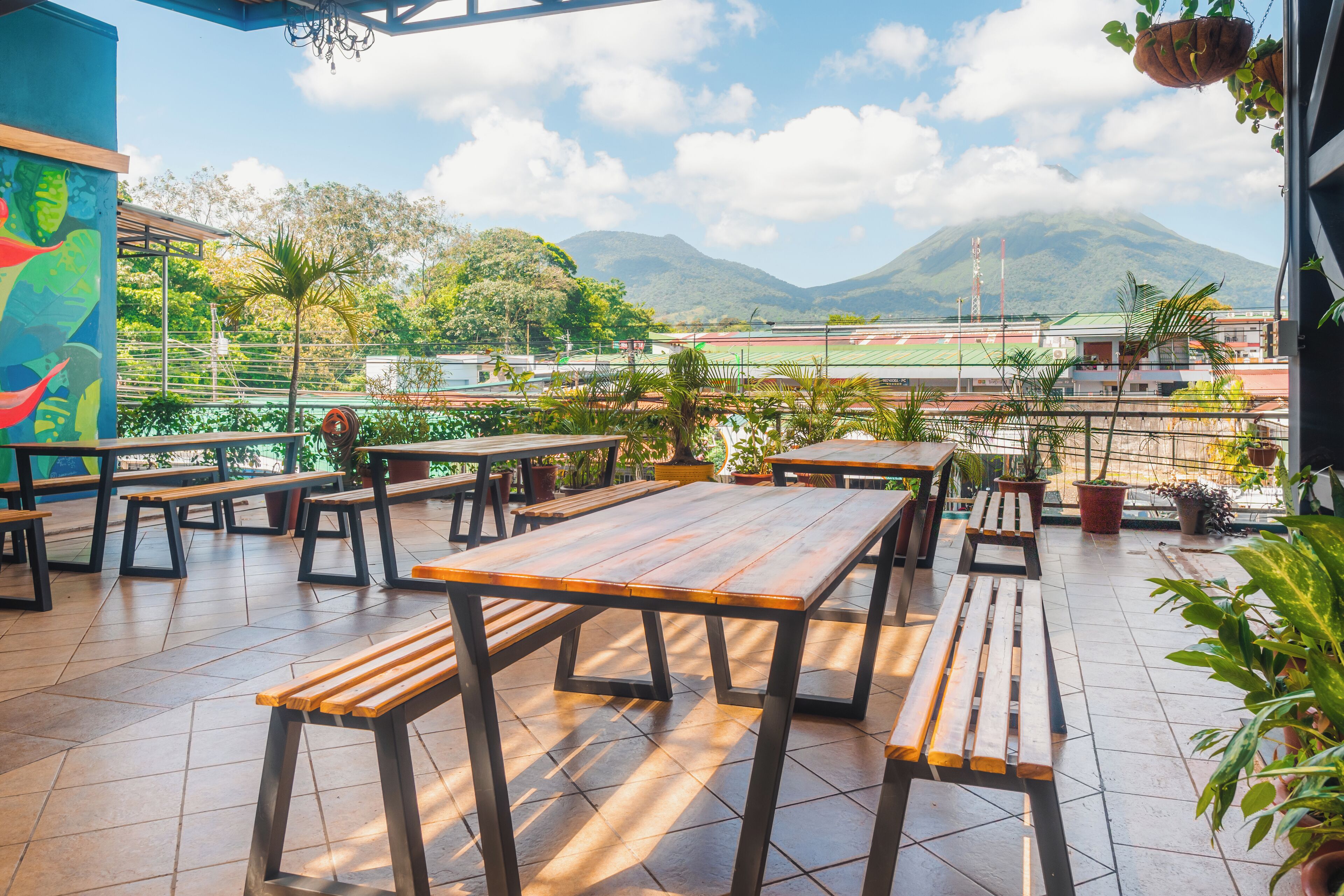 An outdoor patio with wooden tables and benches, surrounded by potted plants and overlooking a sports field and mountains.