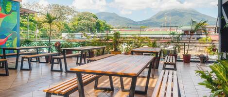 An outdoor patio with wooden tables and benches, surrounded by potted plants and overlooking a sports field and mountains.