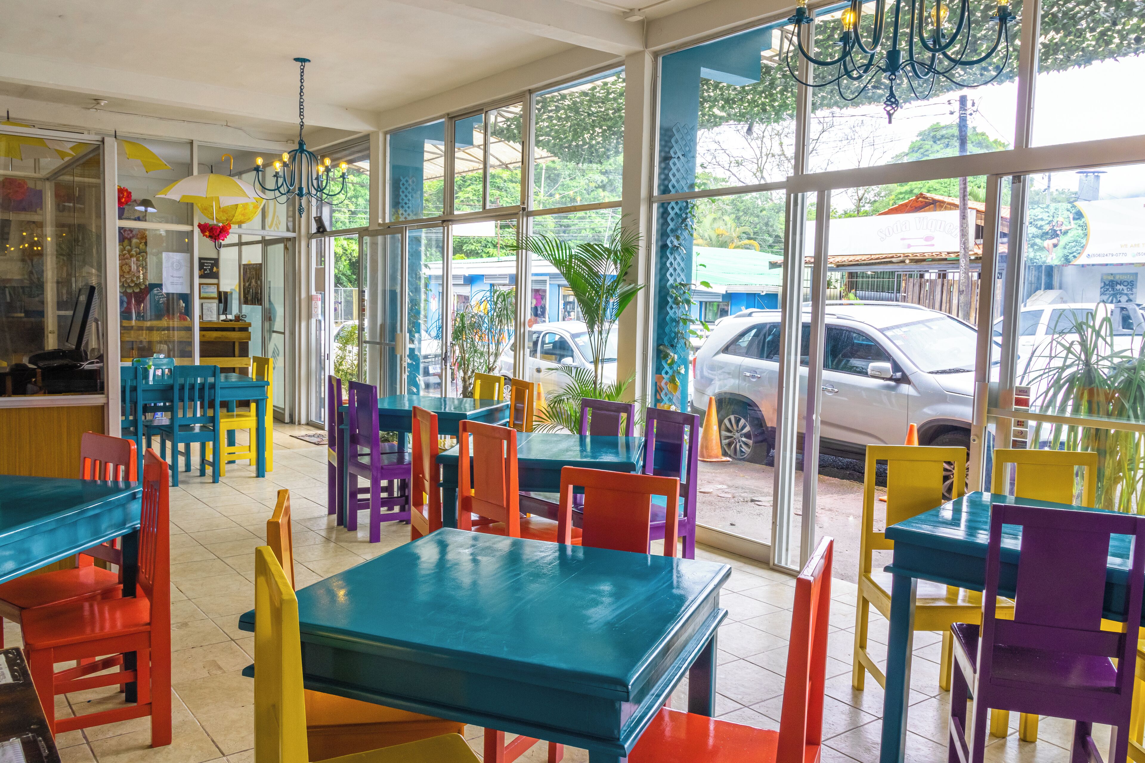 A restaurant with colorful tables and chairs, large windows, and a view of the street outside.