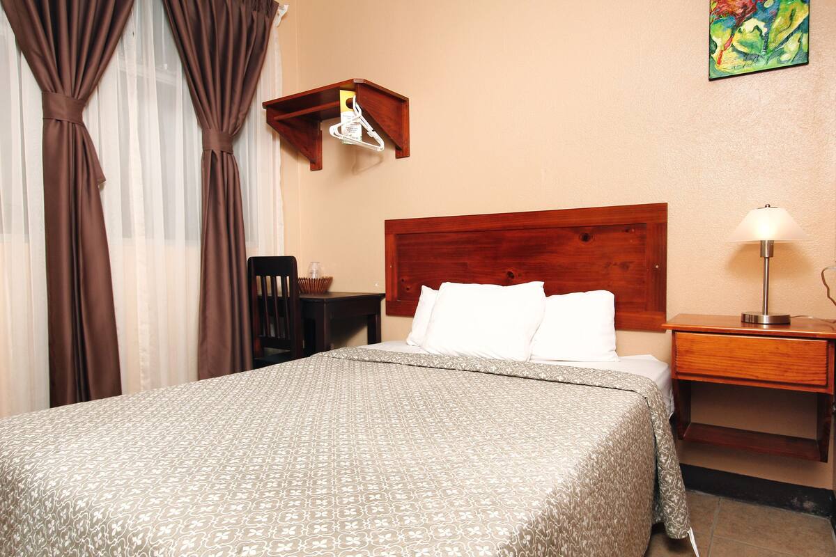 Una cama bien hecha con una colcha estampada, un cabecero de madera, una mesita de noche con una lámpara y un pequeño estante de madera encima de la cama.