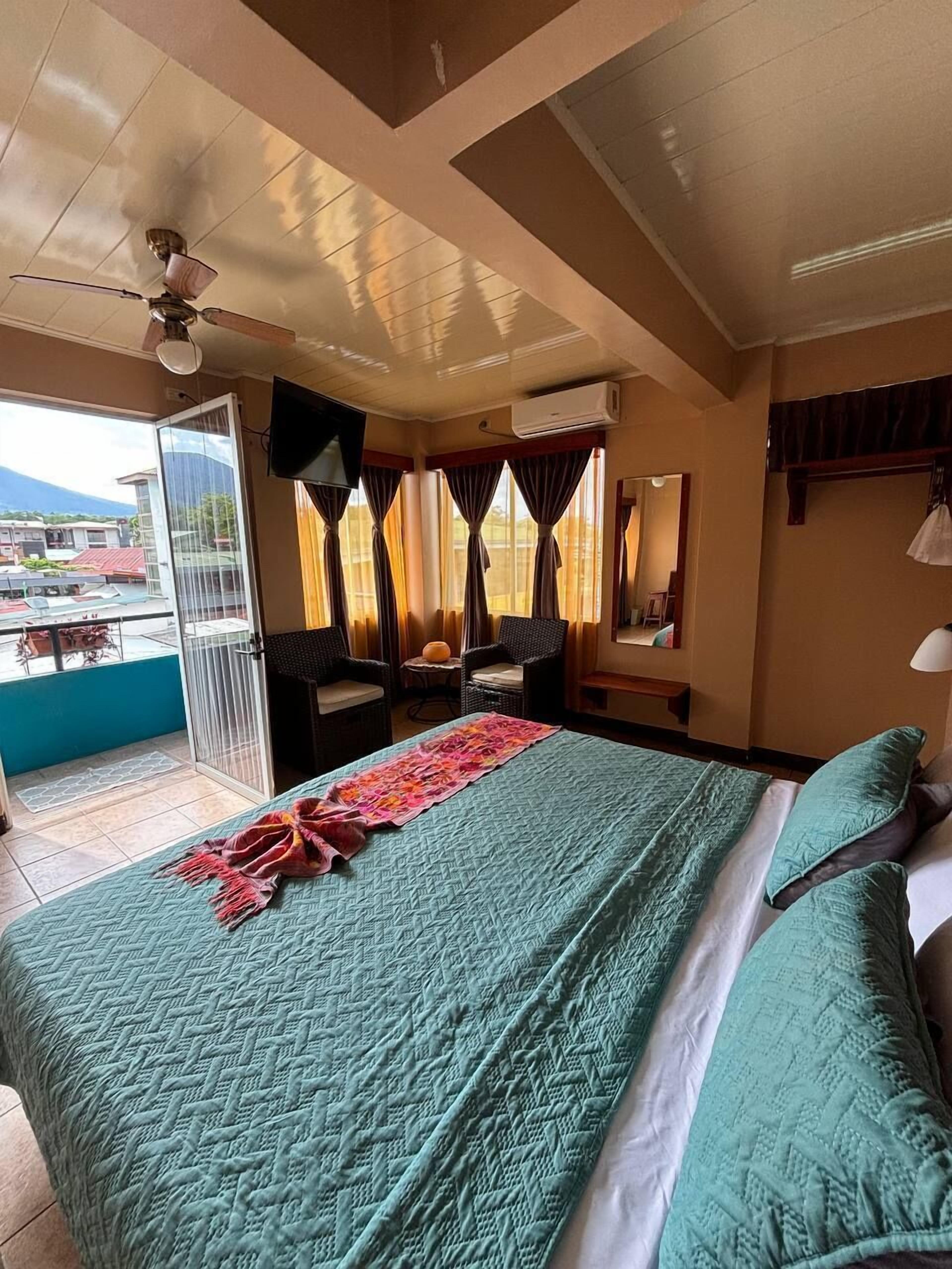 Habitación de hotel con cama, ventilador de techo, televisor y vistas al exterior.