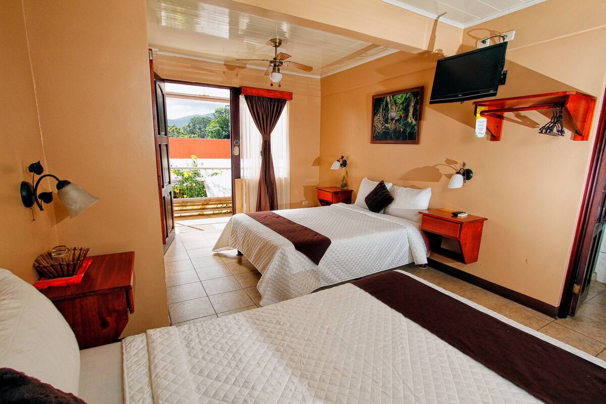 Habitación de hotel con dos camas, ventilador de techo, televisor, un cuadro, mesita de noche y ventana con cortinas.