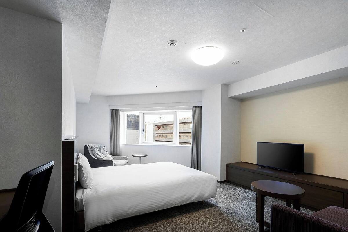 A hotel room with a bed, a TV, a small table, and a window with curtains.