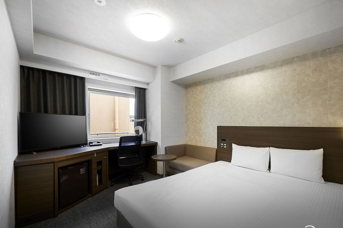 A hotel room with a bed, desk, chair, TV, and a small table.