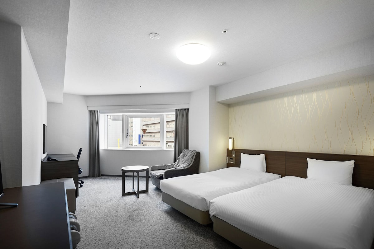 A hotel room with two beds, a desk, a chair, a small table, and a window with curtains.