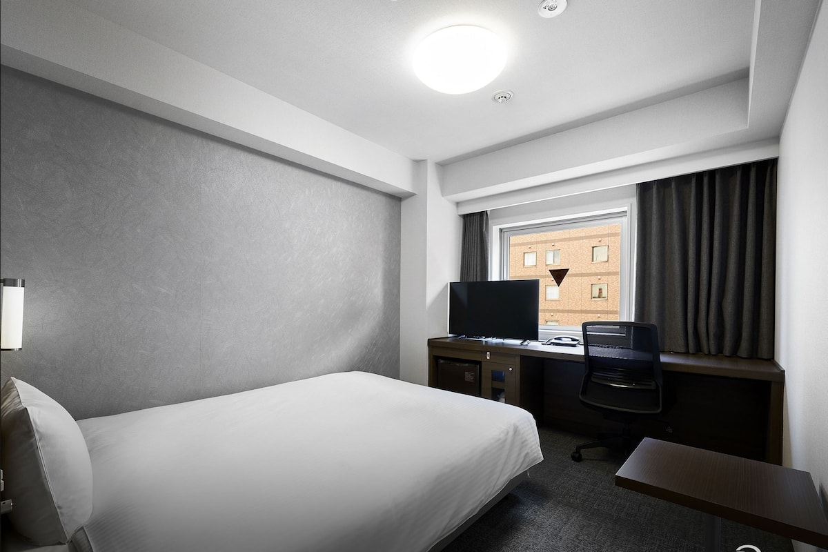 A hotel room with a bed, a desk with a computer, a TV, and a window with curtains.