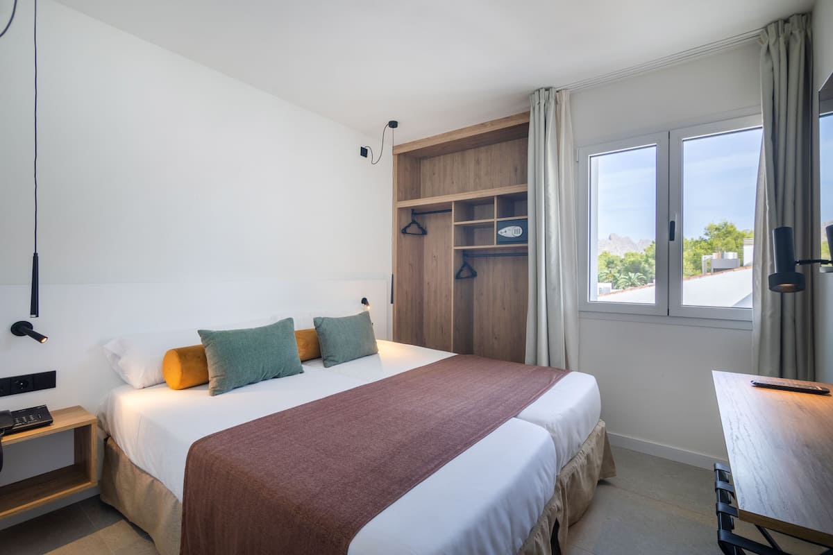 A hotel room with a large bed, a wooden wardrobe, a desk, and a window with a view of greenery and buildings.