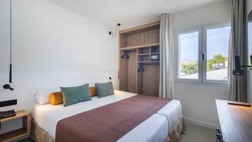 A hotel room with a large bed, a wooden wardrobe, a desk, and a window with a view of greenery and buildings.